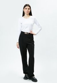 Weißes Langarmoberteil kombiniert mit hochgeschnittenen schwarzen Hosen, die einen goldenen Kettengürtel tragen. Das Model trägt klobige schwarze Schuhe.