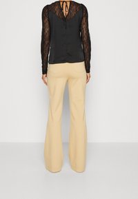 Femme portant un pantalon beige à jambes larges et un haut en dentelle noire à manches longues avec un détail noué au niveau de l'encolure arrière, debout devant un fond uni.