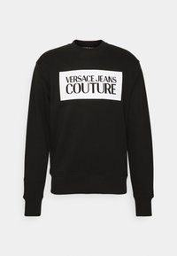 Černá bavlněná mikina s bílým obdélníkovým panelem a nápisem "VERSACE JEANS COUTURE" v tučném černém písmu. Žebrovaný výstřih a manžety.