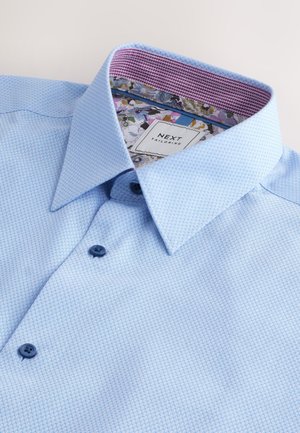 Lyseblå ternet skjortebluse med en struktureret krave, der har mørkeblå knapper og en blomsterprintet inderkrave.