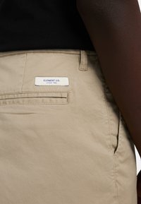 Primer plano de pantalones beige con un bolsillo trasero y una etiqueta blanca que dice "ELEMENT CO. SINCE 1992", usados con una camiseta negra.