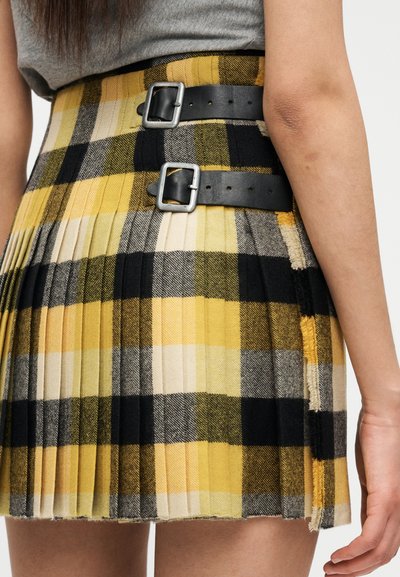 Πλισέ φούστα με κίτρινο, μαύρο και λευκό καρό μοτίβο. Διαθέτει δύο μαύρες δερμάτινες ζώνες με ασημένιες αγκράφες. Λεπτομέρεια με ξεφτισμένες άκρες.