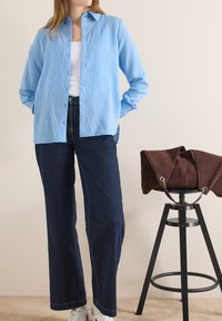 Personne portant une chemise à rayures bleues, un haut blanc, un jean foncé et des baskets blanches, debout à côté d'un tabouret noir sur lequel repose un sac marron.