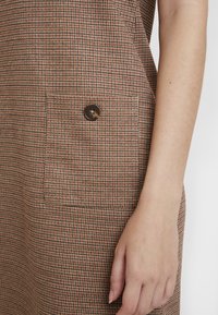 Robe marron en pied-de-poule avec une poche plaquée et un bouton rond marron, présentant un tissu texturé avec une silhouette structurée.