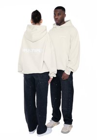 Krémová mikina s logom "MULTIPLY" na chrbte, v kombinácii s čiernymi voľnými džínami. Obe modely majú na sebe tenisky, čím prezentujú uvoľnený vzhľad.