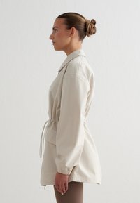 Femme aux cheveux attachés, portant une veste beige clair à taille cintrée et poignets élastiques, debout de profil devant un fond uni.