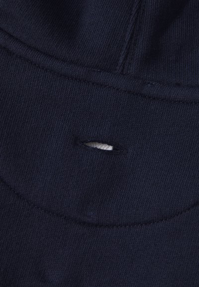 Tummansininen neulottu kangas, jossa pieni vaakasuuntainen repeämä, joka paljastaa alla olevan valkoisen materiaalin, läheltä otettu kuva ompeleesta ja rakenteesta.