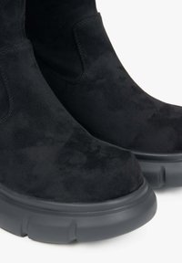 Černé semišové kotníkové boty s kulatou špičkou, elastickými bočními panely a robustní černou gumovou podrážkou. Hladký povrch bez viditelných vzorů.