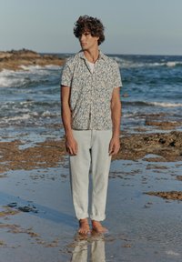 Korte mouwen, knoopshirt met een blauw bladpatroon op een beige achtergrond, gecombineerd met lichtgewassen opgerolde jeans, staand bij het water.