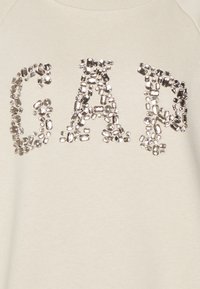 Felpa beige chiaro con decorazioni argentate simili a gemme disposte a formare le lettere "GAP" sul davanti.