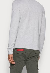 Graues Langarmshirt mit gerippten Bündchen, kombiniert mit olivgrünen Cargo-Hosen, die einen roten "FLYING TIGERS"-Aufnäher und mehrere Taschen aufweisen.