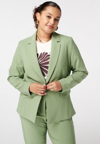 Lichtgroene tailormade blazer met gestructureerde schouders, notch-revers en lange mouwen, gecombineerd met een crèmekleurig shirt met een bruin bloemenpatroon.