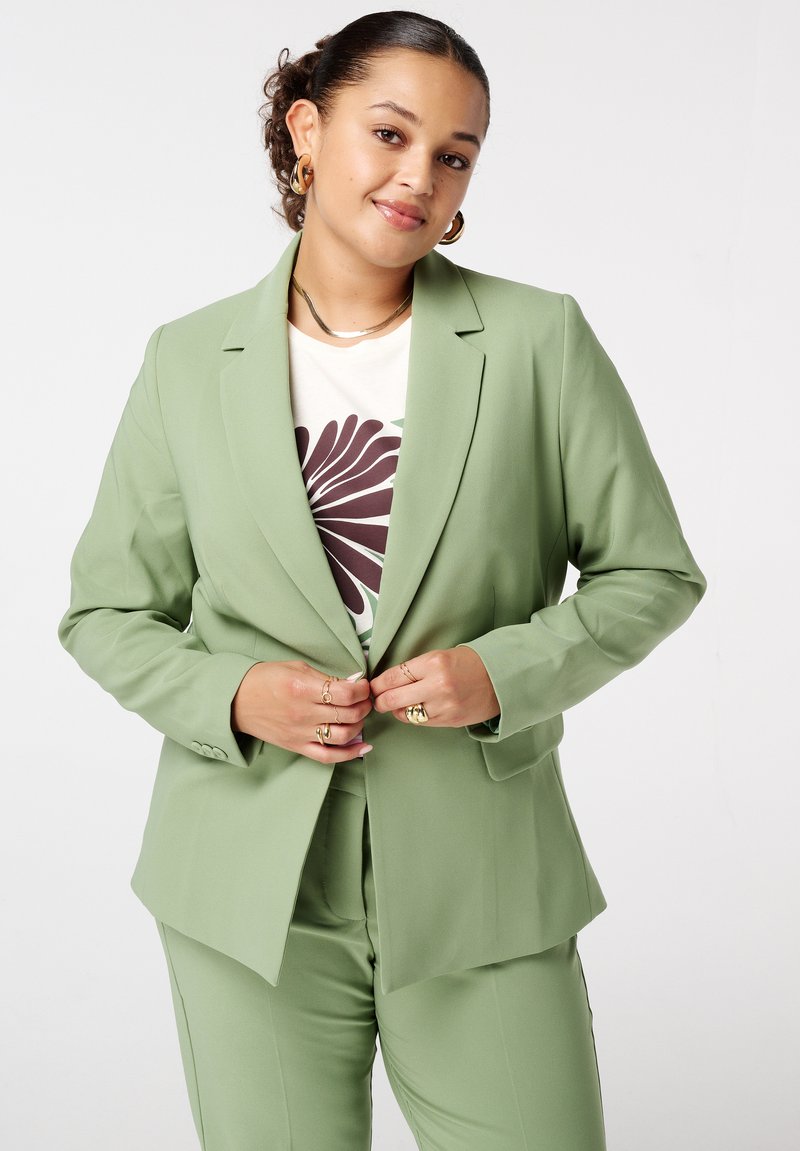 Lichtgroene tailormade blazer met gestructureerde schouders, notch-revers en lange mouwen, gecombineerd met een crèmekleurig shirt met een bruin bloemenpatroon.