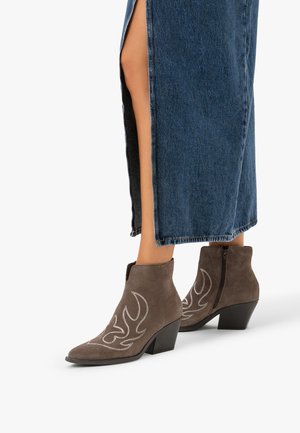 Femme portant des bottines en daim marron avec une couture blanche en forme de flamme et des talons noirs épais, associées à une longue jupe en jean fendue sur le côté.