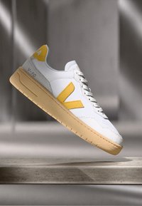 Sneaker bianca con accenti gialli, dotata di una tomaia in pelle texturizzata, lacci piatti e suola in gomma marrone modellata. Design sportivo moderno.