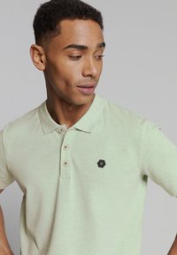Polo shirt in lichtgroen, met korte mouwen, voorzien van een drieknopen sluiting, met een zwart hexagonaal logo op de borst, zachte stofsamenstelling.
