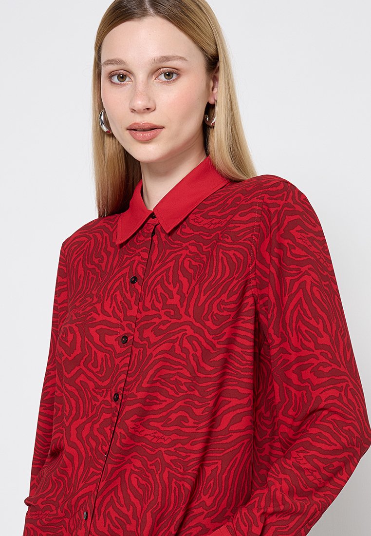 Chemise rouge à boutons avec un motif texturé de rayures de tigre, manches longues, col pointu et boutons noirs sur le devant.