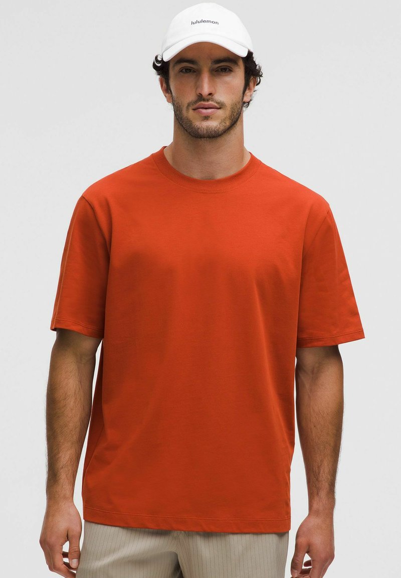 Camiseta de algodón roja con cuello redondo y mangas cortas. Llevada con una gorra blanca que lleva un logo. Diseño simple y casual sin patrones.