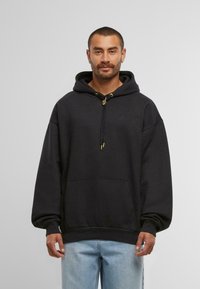 Schwarzer Hoodie mit großer Fronttasche, Kapuze mit Kordelzug und goldenen Akzenten, sowie lässiger Passform. Der Stoff wirkt weich und texturiert.