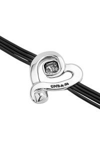Charm plateado en forma de corazón con una piedra preciosa transparente, unido a tres cordones de cuero negro. Presenta grabado "UNO de 50" en el charm.