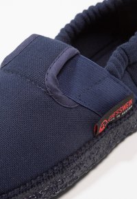 Navyblauwe slipper met een bovenkant van textuurstof, elastische accenten aan de zijkanten en een gestikte donkere zool. Bevat een rood merkembleem.