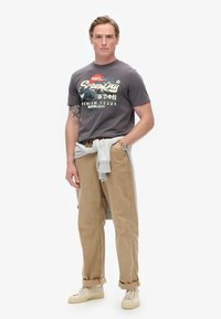 Camiseta gráfica gris con logotipo, pantalones de pana beige y zapatillas crema. Sudadera gris claro atada a la cintura. Mangas cortas y corte relajado.
