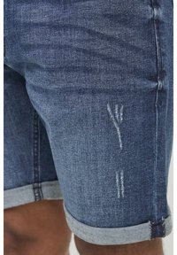 Shorts en denim bleu foncé avec des nuances légères. Présentent un ourlet roulé et des détails déchirés sur le devant. Fabriqués en tissu denim classique.