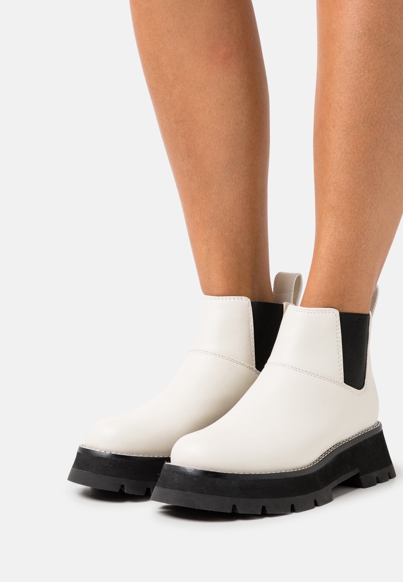 Bottes blanches à cheville avec panneaux latéraux élastiques noirs et semelles épaisses à crampons noires, portées sur des jambes nues sur un fond neutre.