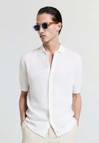 Camicia bianca a maniche corte con bottoni, presenta un motivo a texture e un tessuto lavorato a maglia. Abbinata a occhiali da sole con montatura trasparente e lenti scure.