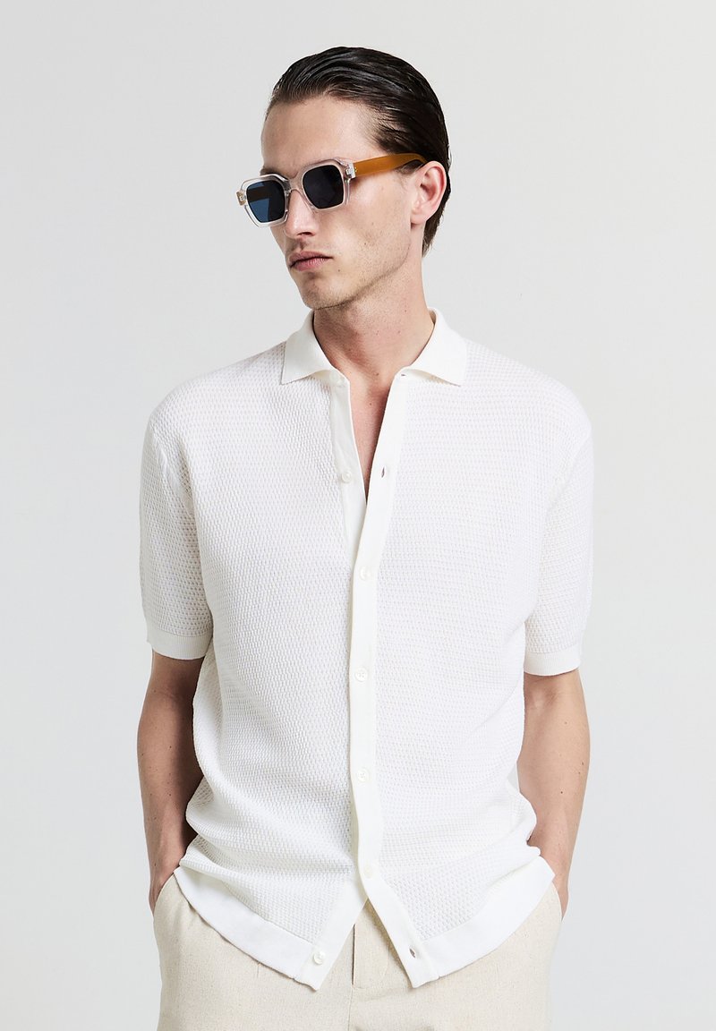 Camicia bianca a maniche corte con bottoni, presenta un motivo a texture e un tessuto lavorato a maglia. Abbinata a occhiali da sole con montatura trasparente e lenti scure.