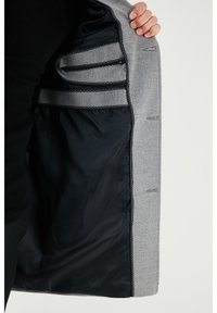 Abrigo de lana gris con forro negro, que presenta múltiples bolsillos y costuras decorativas en los bordes. Abierto para mostrar detalles del diseño interior.