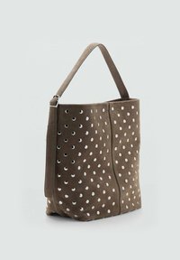 Sac fourre-tout en daim marron avec des découpes circulaires blanches, une sangle plate et une ouverture en haut. Le sac présente une texture lisse et une forme structurée.