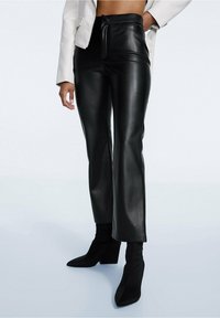 Žena v černých kalhotách s vysokým pasem z umělé kůže, které doplňuje o černé boty na podpatku s špičatou špičkou a zkrácenou bílou bundou.