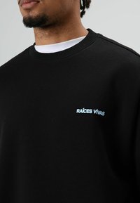 Sweat-shirt noir avec un col rond et des poignets côtelés, portant le texte "RAÍCES VIVAS" brodé en bleu clair sur la poitrine. Texture de tissu douce.