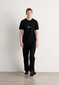 Svart t-skjorte med en liten logo på brystet, kombinert med svarte cargo-bukser. Outfiten har en avslappet passform og flate sko med røde såler.
