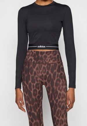 Femme portant un crop top noir à manches longues avec le logo Adidas et des leggings marron taille haute à imprimé léopard.