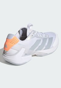 Bílá sportovní bota se šedými pruhy a korálově zbarveným podpatkem. Má texturovaný svršek, šněrování a silnou bílou podrážku.