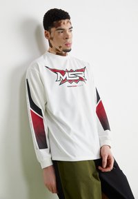 Weißes Langarm-T-Shirt mit einem roten und schwarzen grafischen Logo auf der Vorderseite, ergänzt durch schwarze und rote Designelemente an den Ärmeln.