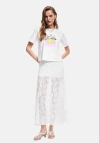 Hvid kort t-shirt med et citronmotiv, parret med en gennemsigtig hvid blonde maxi-nederdel. Modellen har lyse hæle sandaler på.