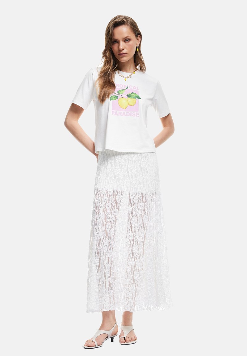 Hvid kort t-shirt med et citronmotiv, parret med en gennemsigtig hvid blonde maxi-nederdel. Modellen har lyse hæle sandaler på.