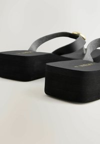 Fekete platform papucs vastag talppal és fekete pántokkal, melyeken kis arany csat látható, világos háttéren bemutatva.