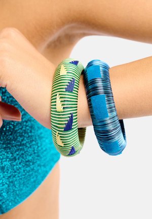 Deux bracelets en tissu à motifs, l'un vert avec des motifs bleus et jaunes, l'autre bleu avec des rayures noires et turquoise, portés au poignet.