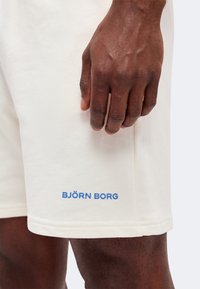 Άτομο με σκούρο δέρμα που φοράει εκρού σορτς με μπλε λογότυπο "BJÖRN BORG", το χέρι ακουμπά δίπλα στο πάνω μέρος του μηρού.