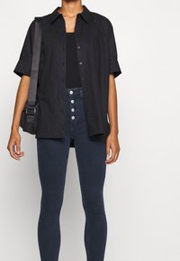 Svart kortärmad skjorta med knappar över en svart linne, kombinerat med mörka denim skinny jeans med fem knapprader.