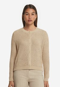 Beige gestrickte Strickjacke mit hexagonalem Muster, rundem Ausschnitt, Knöpfen vorne und gerippten Bündchen. Kombiniert mit hellbeigen Hosen.