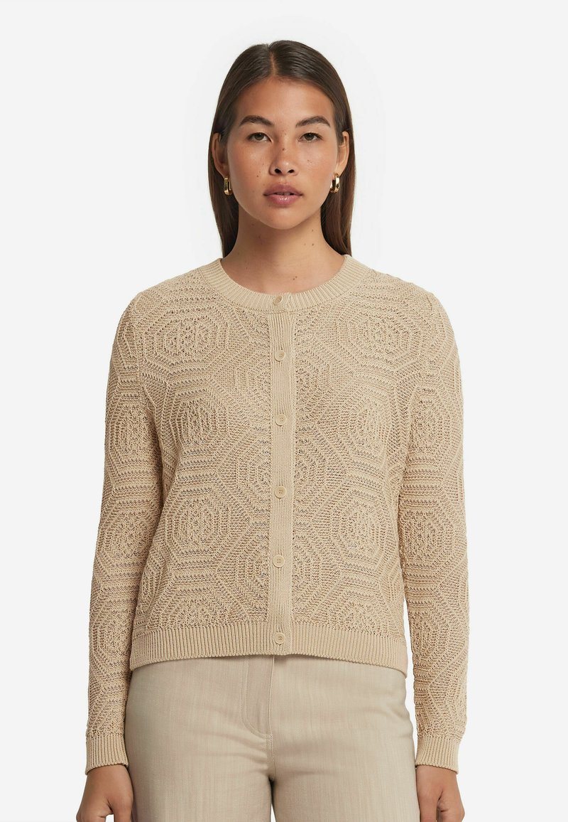 Beige gestrickte Strickjacke mit hexagonalem Muster, rundem Ausschnitt, Knöpfen vorne und gerippten Bündchen. Kombiniert mit hellbeigen Hosen.