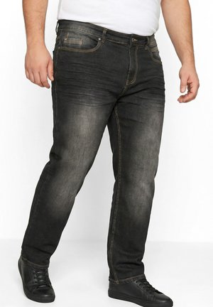 Mann trägt ausgewaschene schwarze Jeans mit kontrastfarbiger Naht und schwarze Schnür-Casual-Schuhe, steht mit leicht gespreizten Beinen.