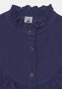 Granatowa koszula z kordurojowego materiału z falbankowym kołnierzykiem, posiadająca dwa czarne guziki z przodu oraz metkę wszytą wewnątrz.