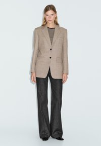 Blazer su misura color marrone chiaro con due tasche frontali a pattina e bottoni neri, abbinato a una maglietta a righe e jeans larghi scuri. Tessuto morbido.