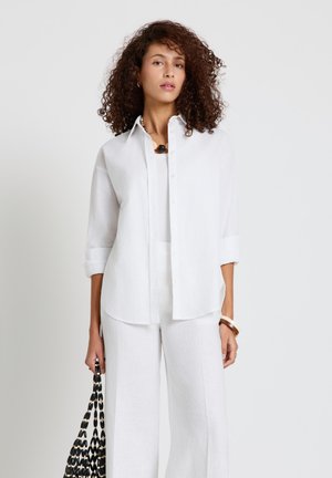 Frau mit lockigem Haar, die ein weißes Hemd mit Knöpfen und weit geschnittene Hosen trägt und eine schwarz-weiße gewebte Tasche an der Seite hält.